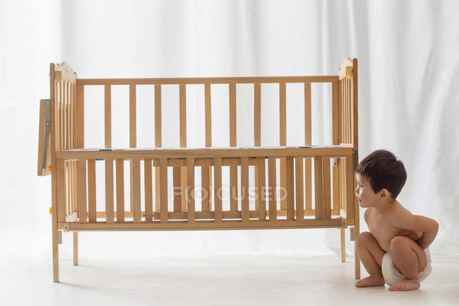 Полный вид восхитительного азиатского малыша в подгузнике, приседающего возле кроватки дома — стоковое фото