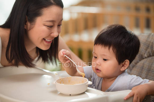 Feliz jovem mãe olhando para adorável criança segurando colher e comer em casa — Fotografia de Stock