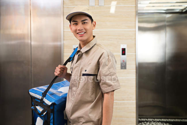 Schöner junger asiatischer Liefermann mit Tasche lächelt in die Kamera im Büro — Stockfoto