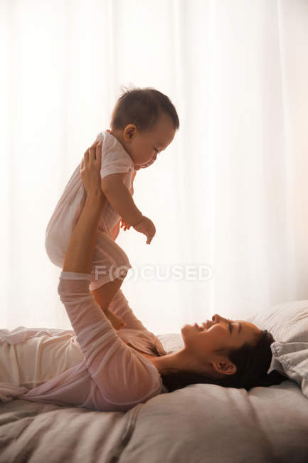 Seitenansicht der glücklichen jungen Mutter, die auf dem Bett liegt und mit dem entzückenden Baby spielt — Stockfoto