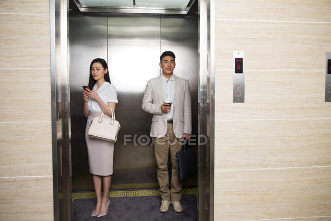 Yougn asiático empresário segurando café para ir e empresária usando smartphone enquanto de pé juntos no elevador — Fotografia de Stock