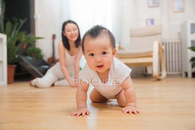 Прелестный азиатский ребенок ползает по полу и смотрит в камеру, счастливая мать сидит позади — стоковое фото