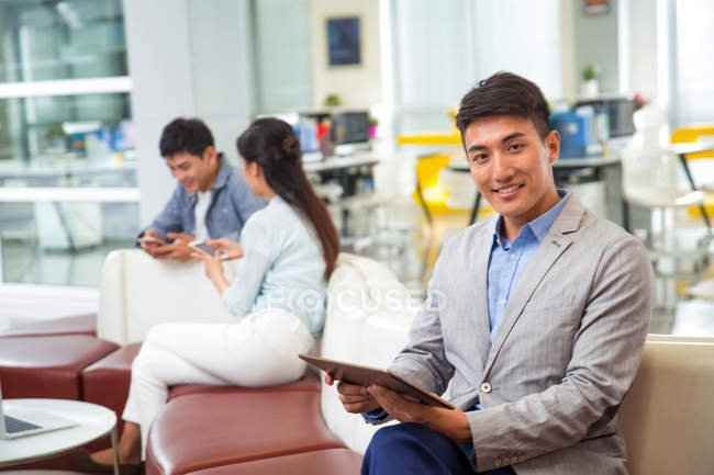 Guapo joven asiático hombre de negocios sosteniendo digital tablet y sonriendo a cámara en oficina - foto de stock