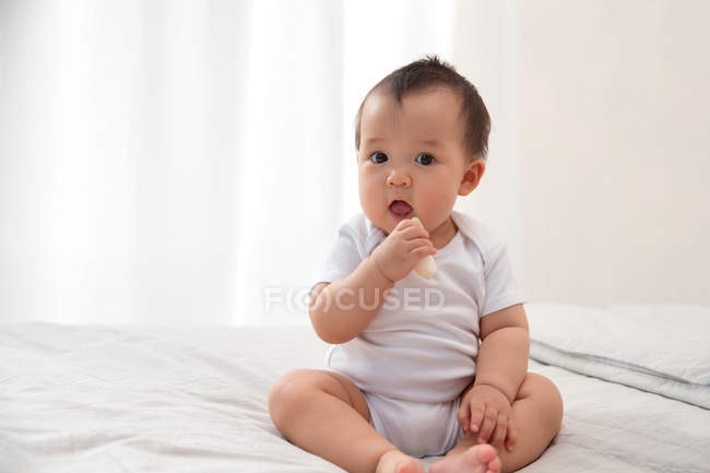 Vorderseite des entzückenden asiatischen Säuglings, der ein Stück geschälte Früchte in der Hand hält und in die Kamera schaut, während er auf dem Bett sitzt — Stockfoto