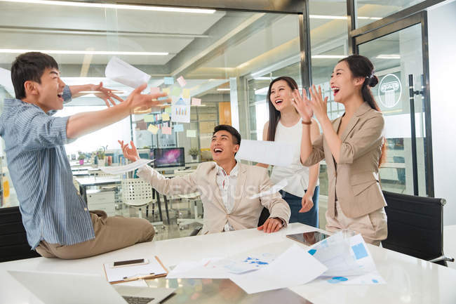 Возбужденные молодые азиатские коллеги по бизнесу бросают газеты в современном офисе — стоковое фото