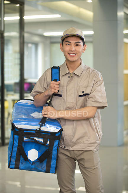 Guapo joven asiático mensajero con bolsa sonriendo a cámara en oficina - foto de stock