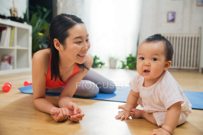 Happy young mother lying on yoga mat and looking at adorable baby at home — Stock Photo