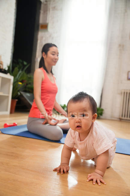 Adorable asian baby crawling on floor while mother meditating behind at home — Stock Photo