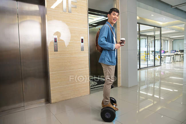 Vue latérale de heureux jeune homme d'affaires asiatique tenant café pour aller et équitation auto-équilibrage scooter près de l'ascenseur — Photo de stock