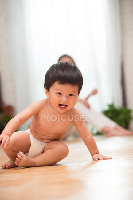 Очаровательный взволнованный азиатский ребенок в подгузнике сидит на полу, счастливая мать сидит позади — стоковое фото