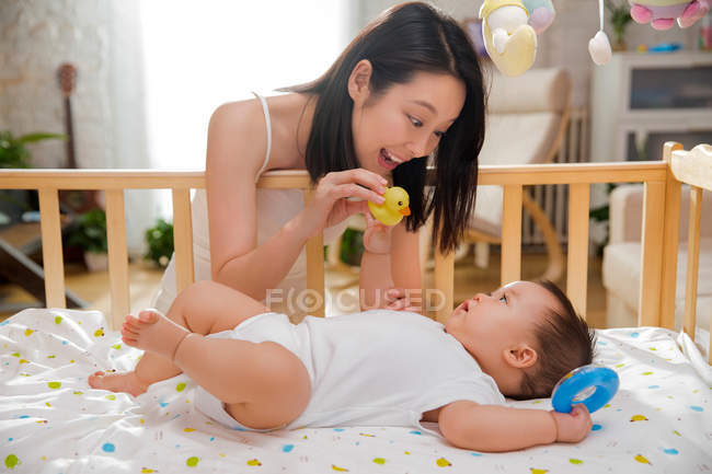 Счастливая молодая азиатская мать, держащая резиновую утку и играя с милым ребенком, лежащим в кроватке — стоковое фото