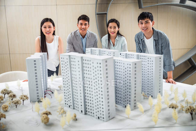 Profissional jovem asiático arquitetos de pé perto do projeto e sorrindo para a câmera no escritório — Fotografia de Stock