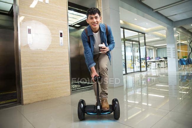 Bello felice giovane uomo d'affari asiatico in possesso di caffè per andare e cavalcare auto-bilanciamento scooter vicino ascensore — Foto stock