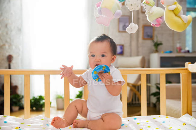 Vue pleine longueur de bébé asiatique adorable tenant jouet bleu et assis dans la crèche — Photo de stock