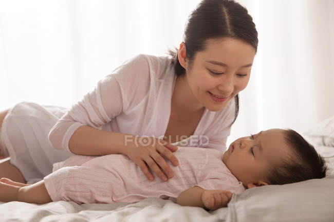 Schöne lächelnde junge Mutter, die das niedliche Baby ansieht, das auf dem Bett schläft — Stockfoto