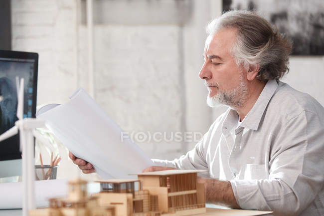 Боковой вид профессионального зрелого архитектора, работающего с blueprint и строительной моделью на рабочем месте — стоковое фото
