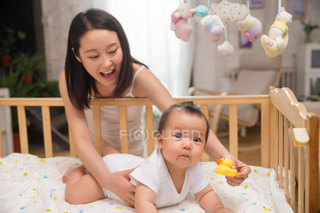 Happy young mother holding rubber duck and playing with cute infant baby lying in crib — Stock Photo