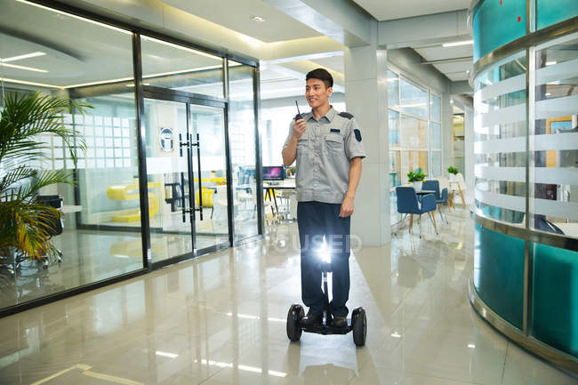 Улыбающийся молодой охранник едет на самобалансирующемся скутере и использует рацию в бизнес-центре — стоковое фото
