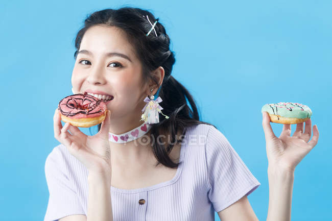 Красива щаслива молода азіатська жінка їсть пончик і посміхається на камеру ізольовано на синьому фоні — стокове фото
