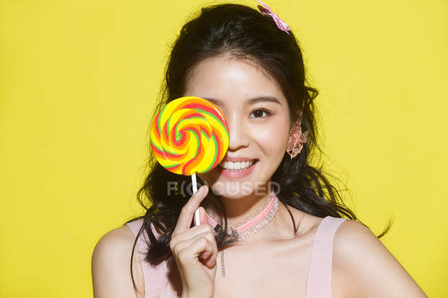 Красива щаслива молода азіатка тримає барвистий льодяник і посміхається на камеру ізольовано на жовтому фоні — стокове фото