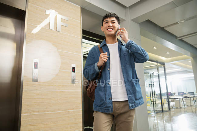 Low angle view of smiling young asian businessman talking by smartphone near elevator in office — Stock Photo