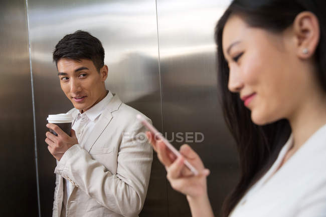 Young businessman holding coffee to go and looking at beautiful businesswoman using smartphone on foreground in elevator — Stock Photo