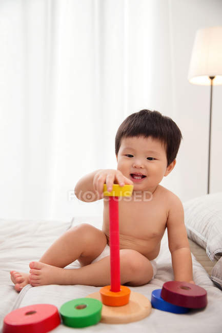 Вид в полный рост восхитительного ребенка в пеленках, сидящего на кровати и играющего с красочной развивающей игрушкой — стоковое фото