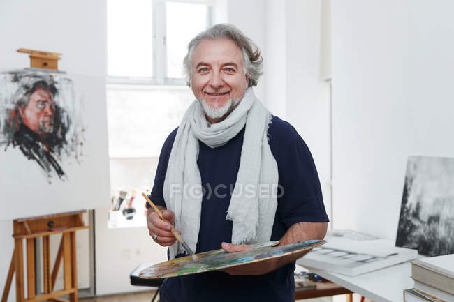 Mature male artist holding palette with paintbrush and smiling at camera in studio — Stock Photo