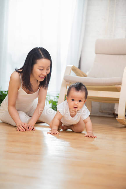 Feliz joven asiático madre mirando su precioso bebé arrastrándose en piso en casa - foto de stock