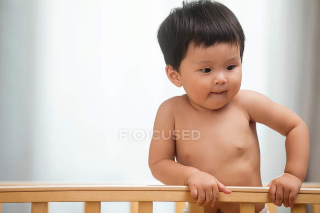 Очаровательный азиатский ребенок в подгузнике, стоящий в кроватке и глядя на дом — стоковое фото