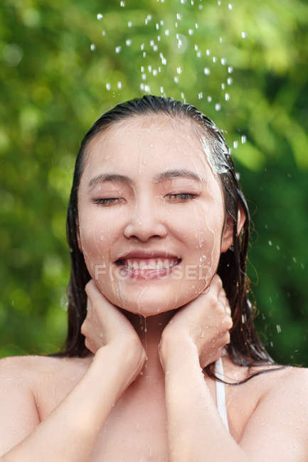 Приваблива усміхнена молода азіатська жінка з закритими очима приймає душ на зеленому природному фоні — стокове фото