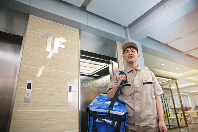Bajo ángulo vista de sonriente joven asiático repartidor hombre con bolsa caminando desde ascensor en centro de negocios - foto de stock