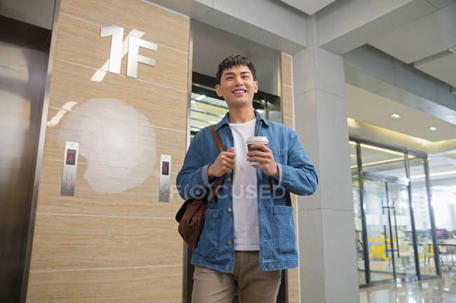Низкий угол зрения улыбающийся молодой азиатский бизнесмен держит кофе идти и ходить возле лифта в офисе — стоковое фото