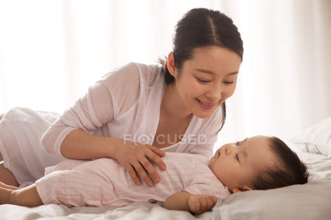 Beautiful happy young asian woman looking at her lovely baby sleeping on bed — Stock Photo