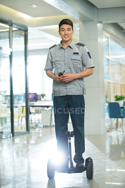 Sorridente giovane guardia di sicurezza cavalcando scooter auto-bilanciamento e utilizzando walkie-talkie nel business center — Foto stock