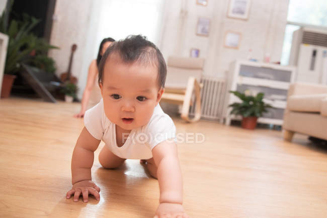 Чарівна азіатська дитина повзає на підлозі, а мати сидить позаду вдома — стокове фото