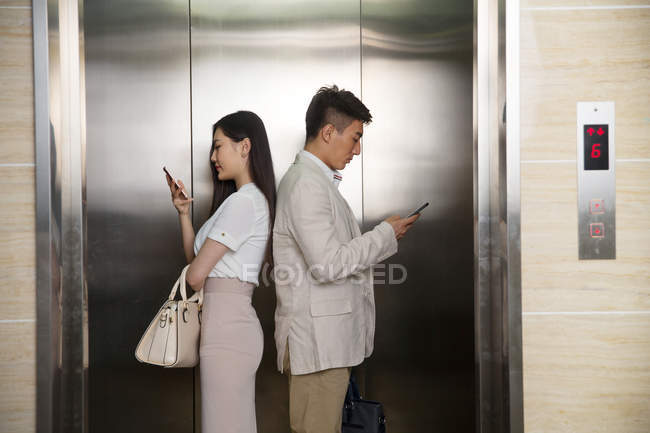 Vista laterale di giovane uomo d'affari asiatico e donna d'affari in piedi back to back e utilizzando smartphone in ascensore — Foto stock