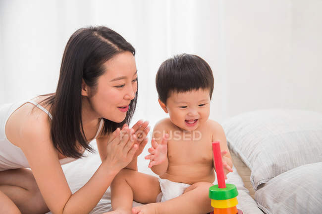 Счастливая молодая мама смотрит на восхитительного ребенка, играющего с разноцветной игрушкой на кровати — стоковое фото