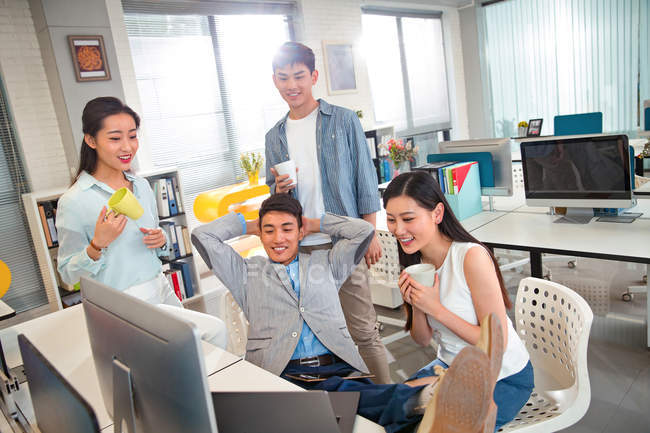 Счастливые молодые профессиональные азиатские бизнесмены, держащие кубки и работающие с компьютерами в офисе — стоковое фото