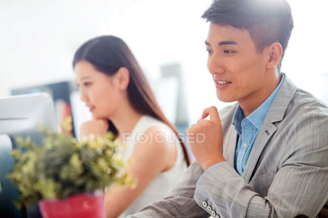 Улыбающийся молодой азиатский бизнесмен, работающий в офисе, красивая деловая женщина, сидящая позади — стоковое фото