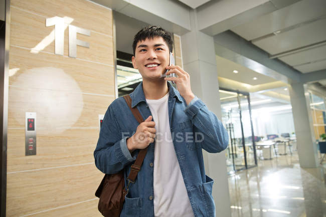 Handsome happy young asian businessman talking by smartphone in office — Stock Photo