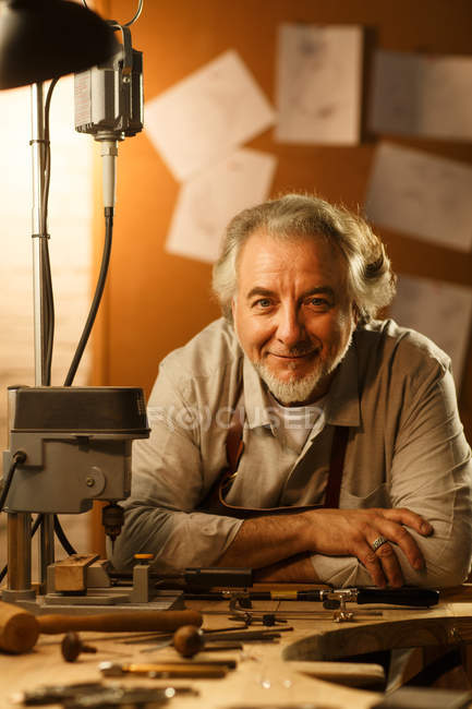 Professional happy mature jewelry designer in apron leaning at table with tools and smiling at camera in workshop — Stock Photo