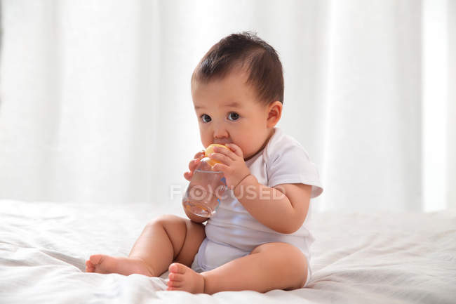 Повний вигляд чарівного азіатського немовляти, що тримає пляшку з водою і сидить на ліжку — стокове фото