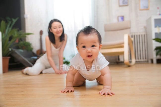 Чарівна азіатська дитина плаче на підлозі і посміхається на камеру, поки щаслива мати сидить позаду вдома — стокове фото