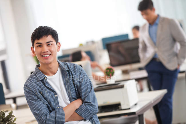 Красивый молодой азиатский бизнесмен со скрещенными руками, улыбающийся в камеру в современном офисе — стоковое фото