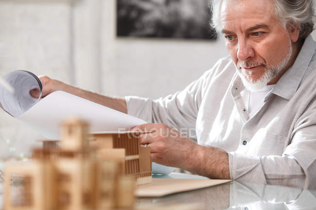 Профессионально сфокусированный зрелый архитектор, работающий с чертежами и моделью здания на рабочем месте — стоковое фото