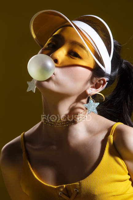 Bela ásia menina no cap e estrela em forma de brincos soprando chiclete e olhando para câmera no estúdio — Fotografia de Stock