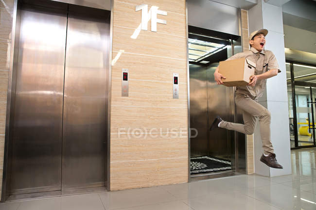Vista de longitud completa del paquete de retención de mensajero masculino joven y corriendo desde el ascensor - foto de stock