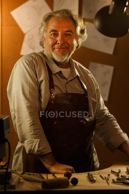 Professionnel mature mâle créateur de bijoux dans tablier debout sur le lieu de travail et souriant à la caméra — Photo de stock