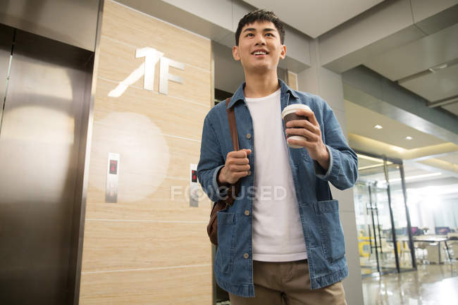 Низький кут зору усміхнений молодий азіатський бізнесмен тримає каву, щоб піти в офіс — стокове фото